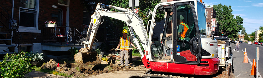 Un professionnel dans un tractopelle et un employé avec une pelle qui réalise une excavation devant un bâtiment résidentiel