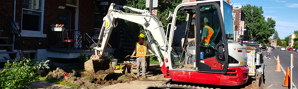 A professional in a backhoe and an employee with a shovel who performs an excavation in front of a residential building